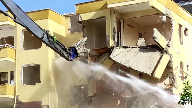 Excavator with hydraulic claw breaks down old multi-storey residential building, water pressure knocks down dust, renovation of housing stock, concept of rebirth and improvement of living conditions