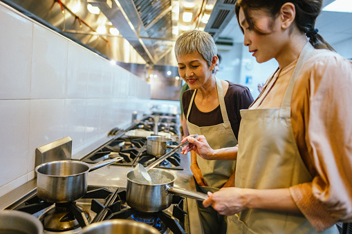 Two Asian Female Adult Students Stirring and Seasoning Ingredients for Dish Prepared During Team Cooking Class in the Kitchen.