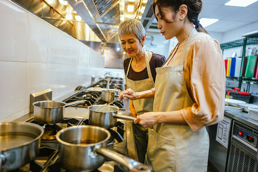 Two Asian Female Adult Students Stirring and Seasoning Ingredients for Dish Prepared During Team Cooking Class in the Kitchen.