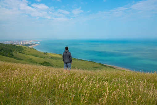 Young man admires beautiful coastal landscape from a viewpoint on top of a cliff. He is on a scenic walk along green cliffs above seaside town of Eastbourne while exploring coast of South England.