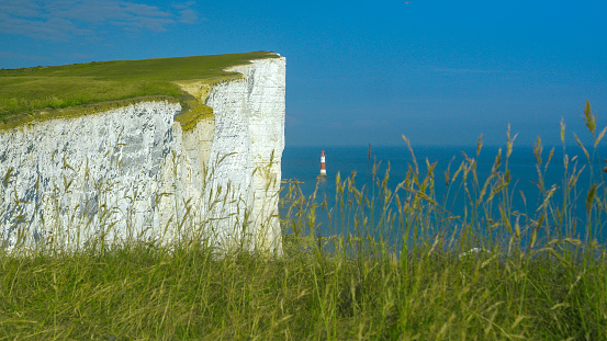 Lighthouse at the end of white walls topped with grass rising above blue sea. Crumbling chalk sea cliffs stretching along picturesque coastline in southern England on a beautiful sunny summer day.