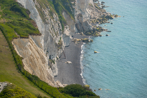 Blue sea gently lapping against rocky shore beneath magnificent white cliffs. Picturesque coastline with chalk sea cliffs topped with grass stretching along ocean in southern England on a sunny day.