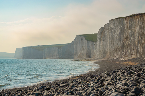Picturesque white cliffs rising above the rolling ocean with a rocky beach below. Tourist attraction with a stunning coastline of chalky sea cliffs covered in green grass next to the English Channel.