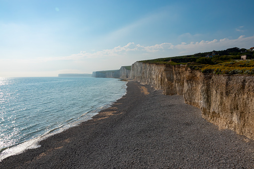 Small ocean waves rolling onto the rocky beach beneath magnificent chalk cliffs. Tourist attraction with a stunning coastline of chalky sea cliffs covered in green grass next to the English Channel.