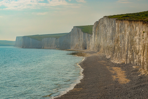 Rocky beach under majestic white cliffs that rise above the blue wavy ocean. A tourist location with a beautiful sea coast of chalky sea cliffs covered with green grass, next to the English Channel.