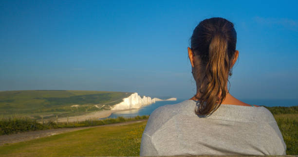 CLOSE UP, DOF: Back view of young tourist admiring stunning Seven Sisters cliffs rising above blue sea. Female holidaymaker is exploring coastal part of South England famous for breathtaking scenery.