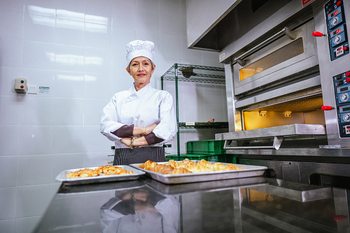 An Asian senior woman baker is looking at the camera. She is dressed in chef attire, including a chef's dress and hat, and is cooking in the kitchen. Together with another chef, they are baking. She is seen taking freshly baked cookies out of a modern electric oven in the kitchen.