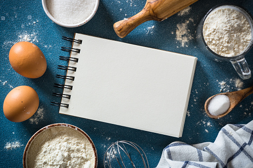 Baking background with cooking book and ingredients. Flour, sugar, eggs and utensil at light stone table. Top view with copy space.