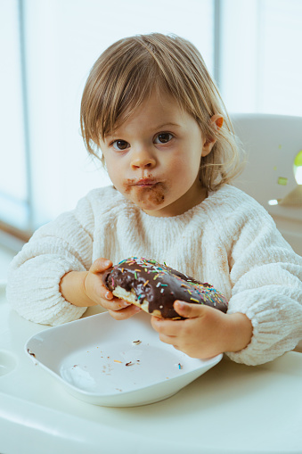 Cute toddler eats donuts with chocolate, harm from sugar to the health of a child, tooth problems, dentist for kid. Messy eating of sweets