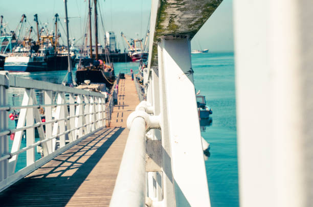 Ko This image offers a unique perspective from a harbor walkway, leading the eye towards a fleet of moored vessels. The white railing creates a striking visual guide, contrasting with the wooden planks underfoot and the tranquil blue sea beyond. The composition captures the essence of a coastal journey, filled with the anticipation of maritime adventure. Perfect for themes related to travel, nautical exploration, and the beauty of seaside destinations.