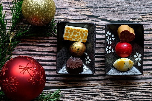 Two small plates with 3 pieces of delicious homemade chocolate on each, on a wooden, rustic background with copy space around. Christmas balls and spruce green on the left side of the plates