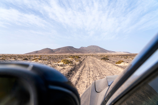 The image captures beauty of Jandia, Fuerteventura, in the Canary Islands, as seen from the driver's point of view. A rental car, its tires covered in dust and sand, traverses a winding off-road path amidst the vast expanse of golden dunes and volcanic terrain.