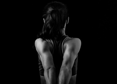 Strong sporty woman doing the arms and shoulders stretching exercise holding the hands in fist behind the back to relaxing back side of body in sport wear. Closeup black and white