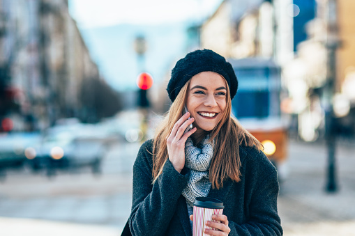 Smiling young woman on street using smartphone. Beautiful woman talking on the phone in urban environment. Happy woman having call on a winter day outdoor in the city