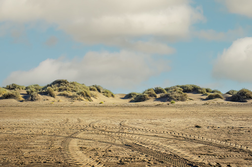 sandy landscape with tire tracks imprinted on it. In the background, there are sand dunes covered with some vegetation under a partly cloudy sky.