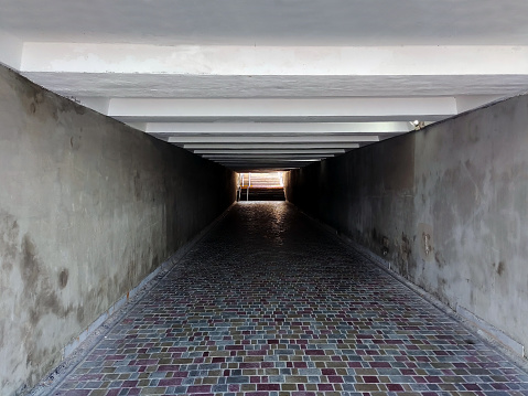 Underpass with tiled floor in perspective with concrete walls. At the end of the transition, the stairs up are illuminated by the sun
