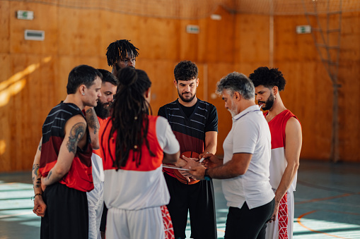 An interracial team is working with their coach on game tactics on their training on court.Senior basketball trainer is explaining game plan and writing on clipboard while interracial team is watching