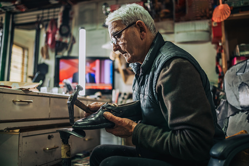 Profile view of an old handyman repairing shoes at shoemaker workshop. A senior cobbler is sitting at his small workshop and fixing shoes and using hammer. A craftsman is repairing shoes at workshop.