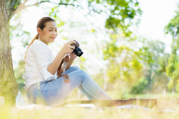 The Asian woman is enjoying taking photos and relaxing under the tree. The Asian woman is looking at the photos on the digital camera.