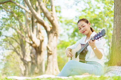Asian beautiful woman sits under the tree at the park and enjoy playing an acoustic guitar. Woman relaxing at the park sitting on the glass field and playing a guitar.
