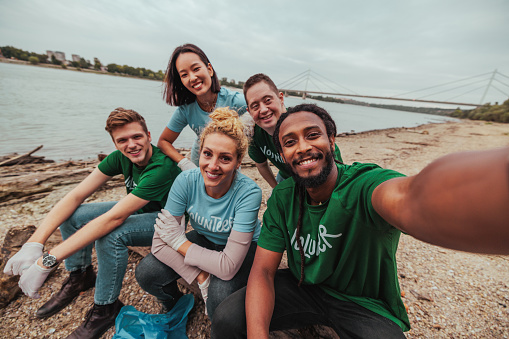 Group of inclusive, Caucasian, African-American, down syndrome, male and female, multiethnic volunteers in uniforms are sitting on a beach taking a selfie after collecting garbage