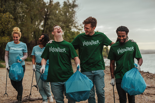 Three male volunteers in uniforms, one Caucasian, one African-American and one with down syndrome are walking by the beach and laughing out loud, collecting trash and talking while their colleagues follow