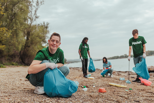 Guy with Down syndrome smiling and collecting plastic in a plastic bag by the river on earth day with his team