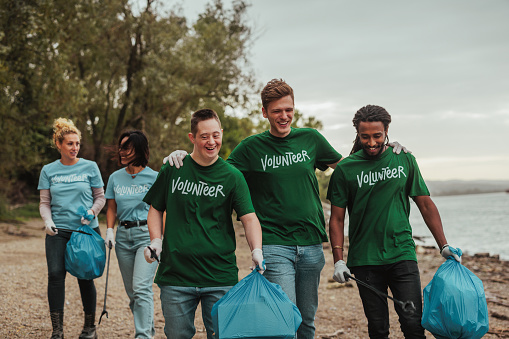 Three male volunteers in uniforms, one Caucasian, one African-American and one with down syndrome are walking by the beach and talking, collecting trash while their colleagues follow