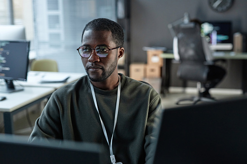 Minimal portrait of focused Black man using computer in IT development office with code lines reflection in glasses copy space