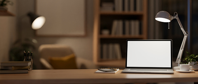 A modern, cozy home office at night with a white-screen laptop computer mockup, a coffee cup, and low light from a table lamp on the table. 3d render, 3d illustration