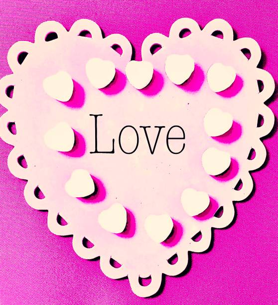 A light pink paper heart embellished with scalloped edges features the typed word “Love” surrounded by white heart-shaped hard candies on a hot pink paper background for Valentine’s Day.