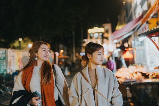 In the photograph, two young asian women are captured taking a nighttime stroll on a vibrant street in Cheung Chau, Hong Kong
