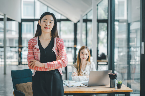 Business team analytics concept, A confident young woman standing arms crossed with her colleague in the background in a modern, light-filled office environment.