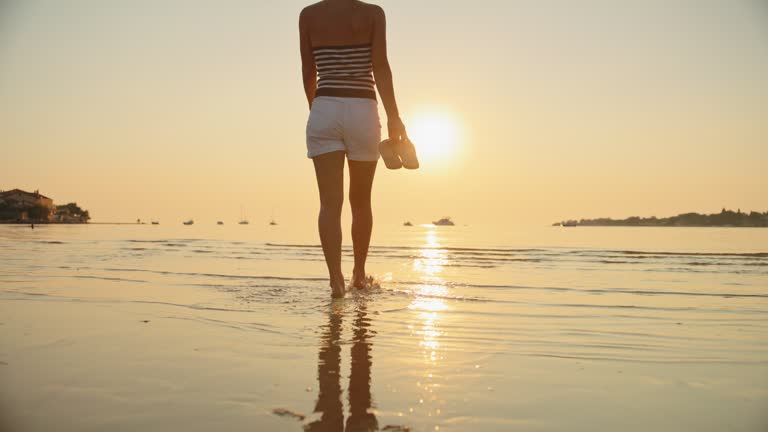 SLO MO Shot of Woman Holding Slippers Walking on Shore at Beach During Sunset