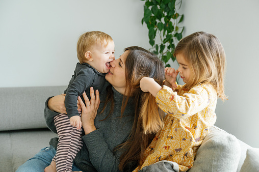 A candid moment captures a mother laughing as her toddler playfully licks her face, with the older sibling joining in the fun, family concept, copy space for text, logo