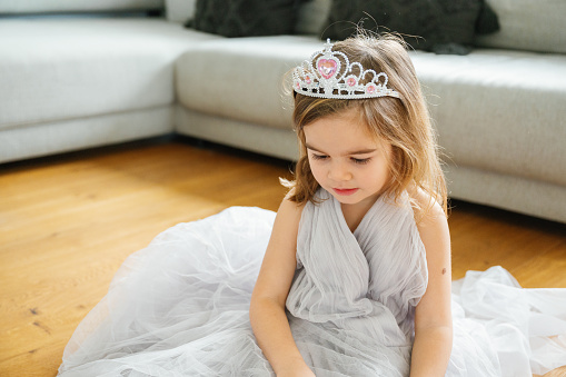 An adorable girl smiles in a tulle dress, with a tiara, sitting on floor, wearing long gray tulle dress joy and princess vibes at home