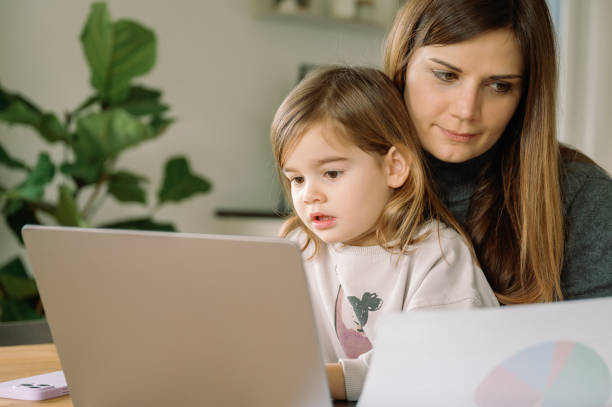 istock young freelancer woman with daughter at home. Self-employed mother working from home while daughter is sitting in her lap and looking laptop 2014419434