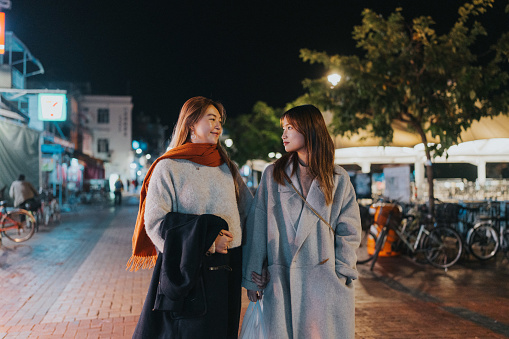 On a pleasant evening, two youthful Asian women stroll through the streets of Cheung Chau, Hong Kong, while the alluring scent of a nearby Dai Pai Dong  fills the air