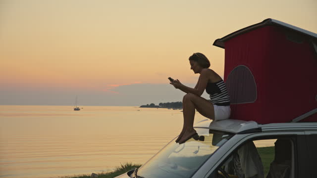 SLO MO Shot of Woman Using Mobile Phone While Sitting on Top of Camping Van at Sunset