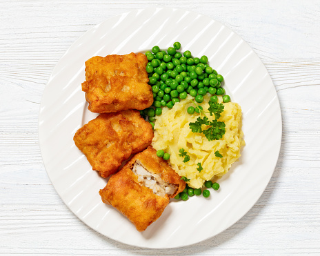 crispy fried fish fillet with mashed potatoes and green peas on white plate on wooden table, horizontal view from above, flat lay, close-up