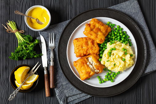 crispy fried fish fillet with mashed potatoes and green peas on plate on black wooden table with rustic cutlery, mustard, lemon, horizontal view from above, flat lay