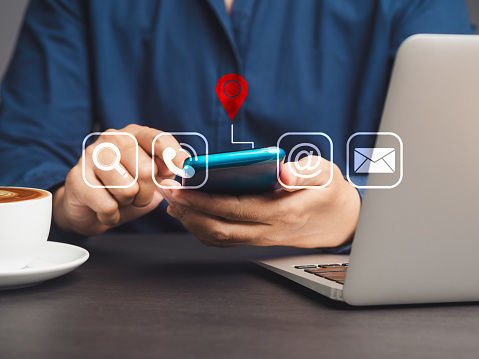 Contact us. Businessman holding a smartphone with a location red pin symbol, search, email, telephone, and address icons while sitting at a desk. Customer service call center.