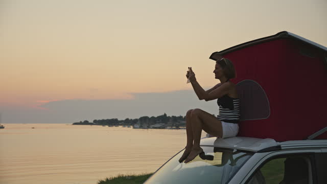 SLO MO Shot of Woman Using Mobile Phone While Sitting by Tent on Top of Van at Sunset