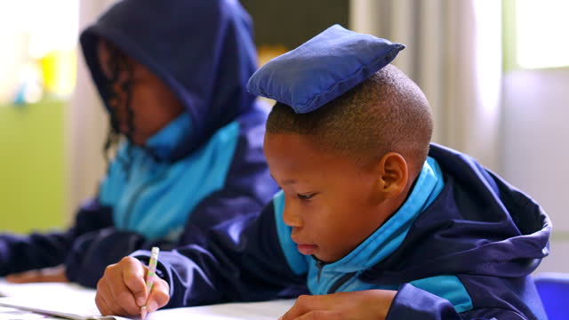 Child, classroom and writing in book at desk with bean bag on head or education, concentration or discipline. Students, school and pencil at South Africa academy for learning, teaching or knowledge
