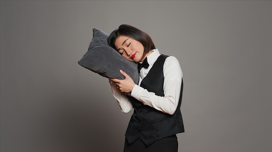Asian front desk staff falling asleep with pillow in hand, feeling extremely tired and overworked. Receptionist napping over grey background, exhausted concierge with fatigue. Camera A.