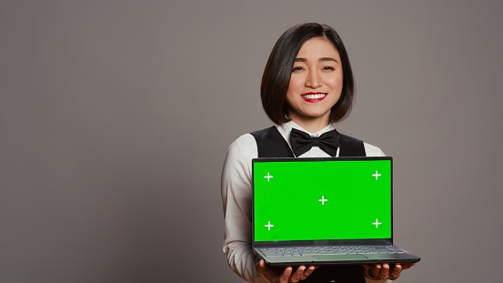 Asian hotel concierge holding pc with greenscreen on camera, presenting isolated mockup template on screen. Receptionist showing laptop with blank chromakey layout on display. Camera B.
