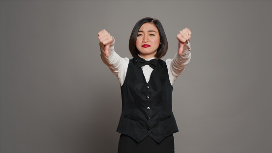 Receptionist showing thumbs down sign over grey background, expressing negativity and disapproval in studio. Woman hotel concierge giving dislike symbol, feeling displeased. Camera A.