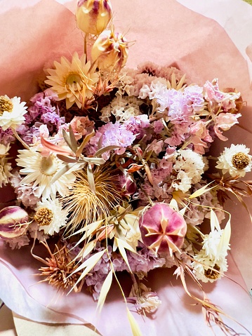 Vertical high angle closeup photo of a variety of dried flowers arranged in a posy wrapped around with pale pink tissue paper, on display for sale in a Florist shop.