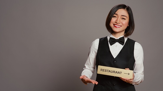 Asian hotel concierge holding restaurant sign to indicate direction, pointing towards dining area. Receptionist assisting clients to enjoy all amenities, stands over grey background. Camera B.