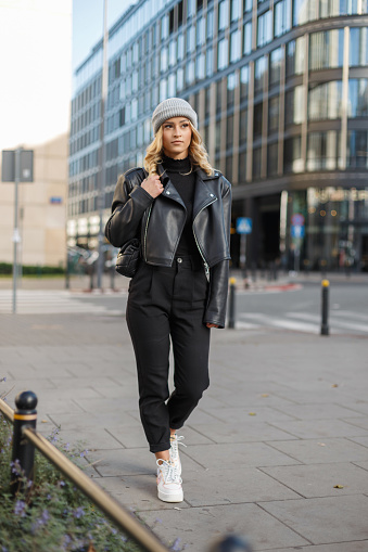 Beautiful fashion young girl with a knitted hat in black clothes with a leather jacket, sweater, trousers, white sneakers and a bag is walking in the city